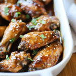 Air fryer teriyaki chicken wings in a white baking dish, coated in a sticky sauce and sprinkled with sesame seeds and fresh herbs.